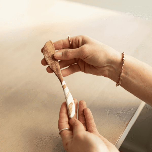 Hand holding a Lynn & Liana acacia wood cheese spreader with a white and gold Gold Quartz resin handle