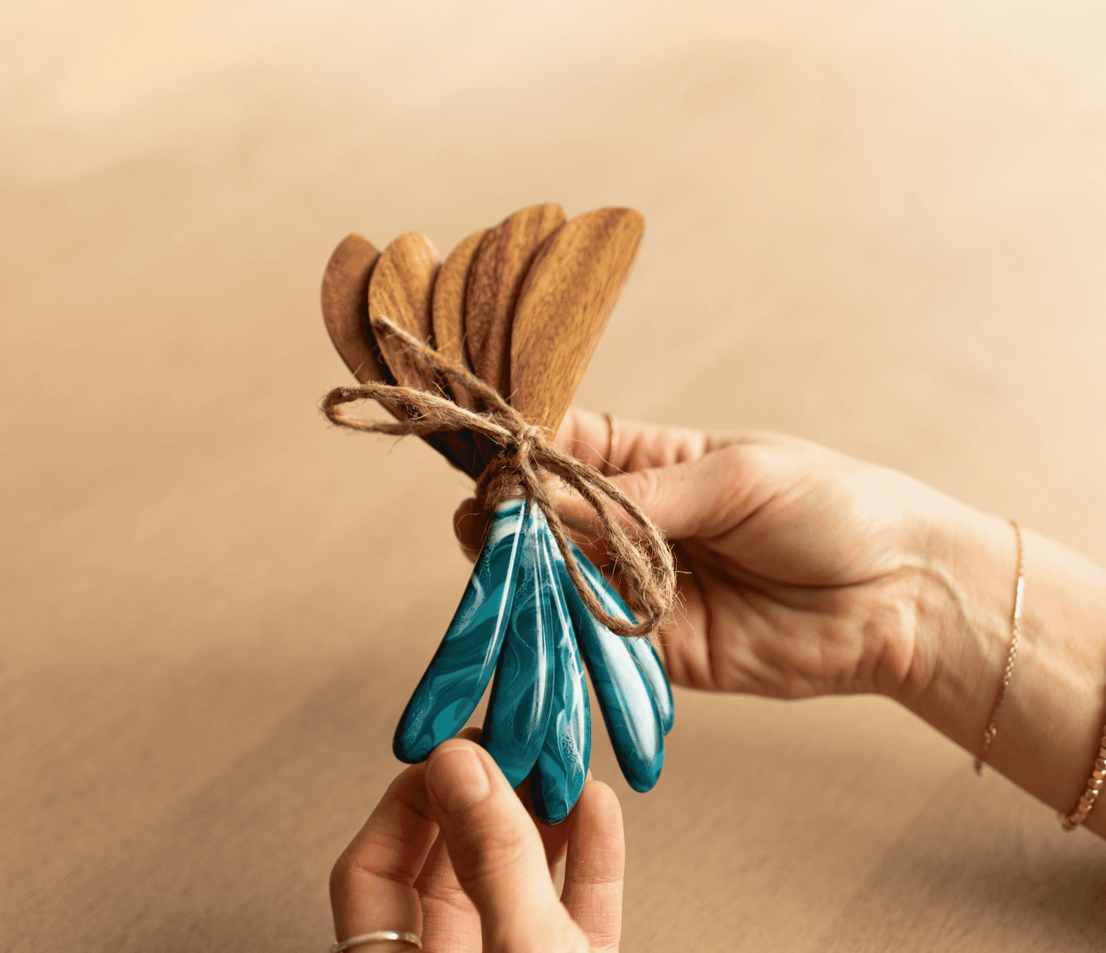 A hand holding a bundle of five Lynn & Liana acacia wood cheese spreaders with teal Ocean Vibes resin handles, tied with twine