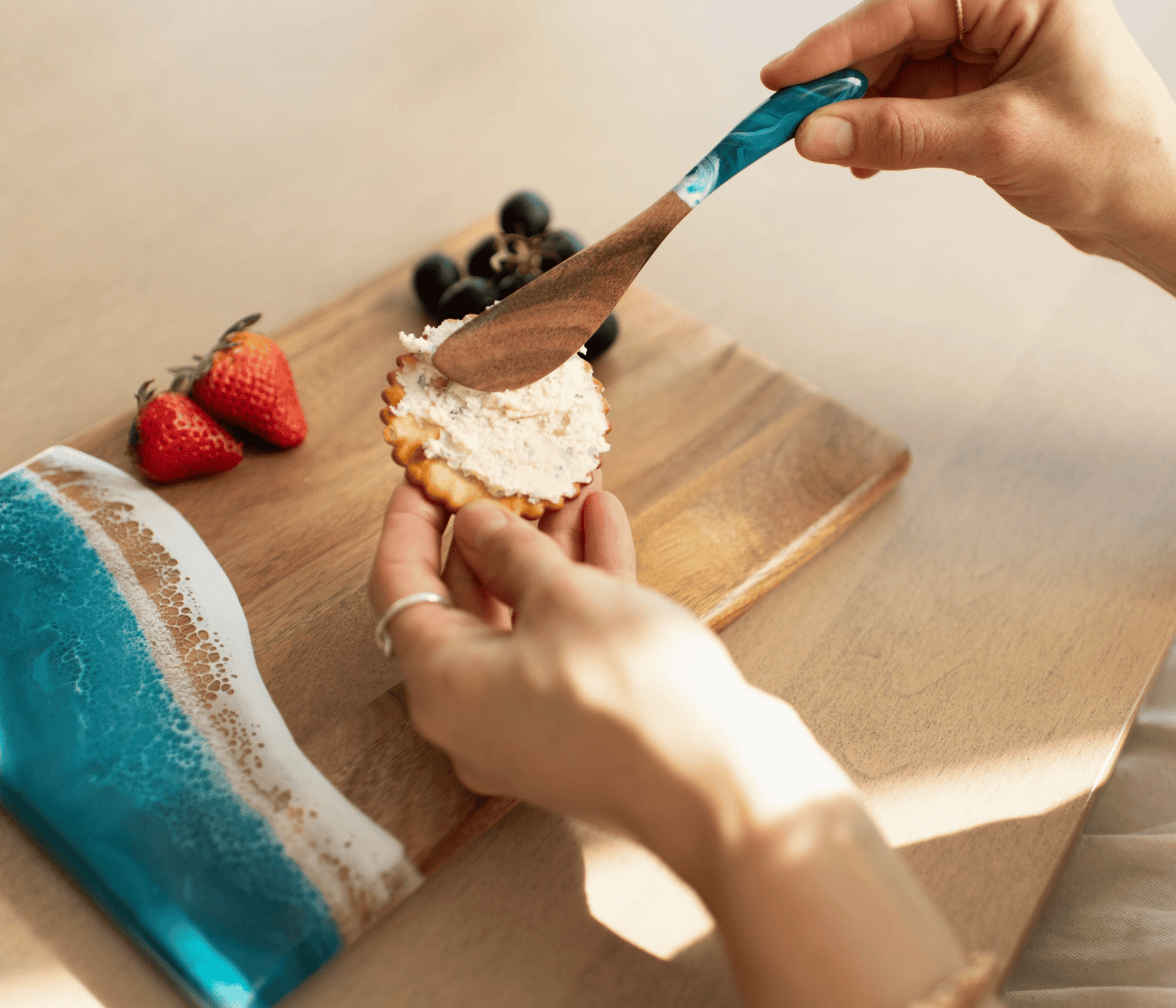 Teal Ocean Vibes acacia cheese spreader spreading soft cheese onto a cracker on a charcuterie board with strawberries and grapes