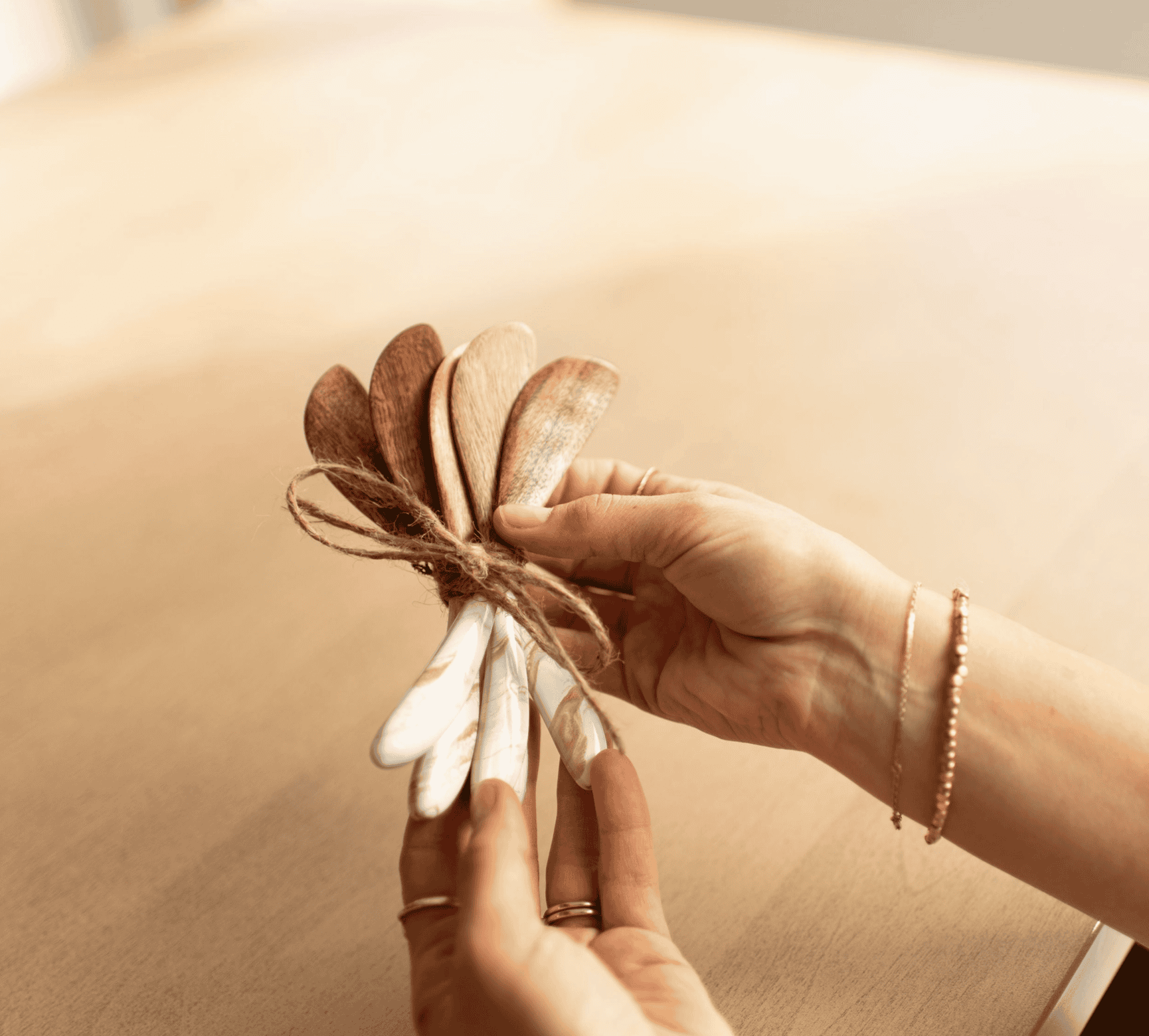 A hand holding a bundle of Lynn & Liana acacia wood cheese spreaders with white and gold Gold Quartz resin handles, tied with twine
