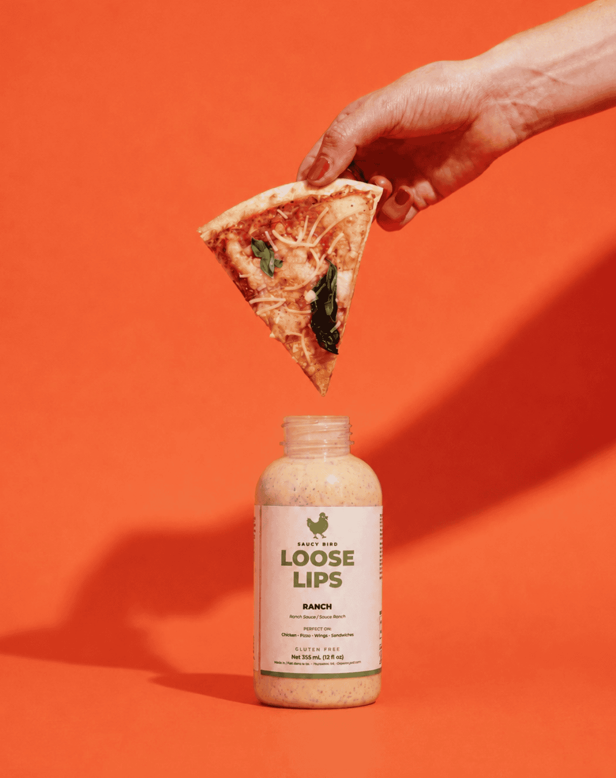 A slice of pizza held above an open bottle of Saucy Bird Loose Lips Ranch Sauce on an orange background