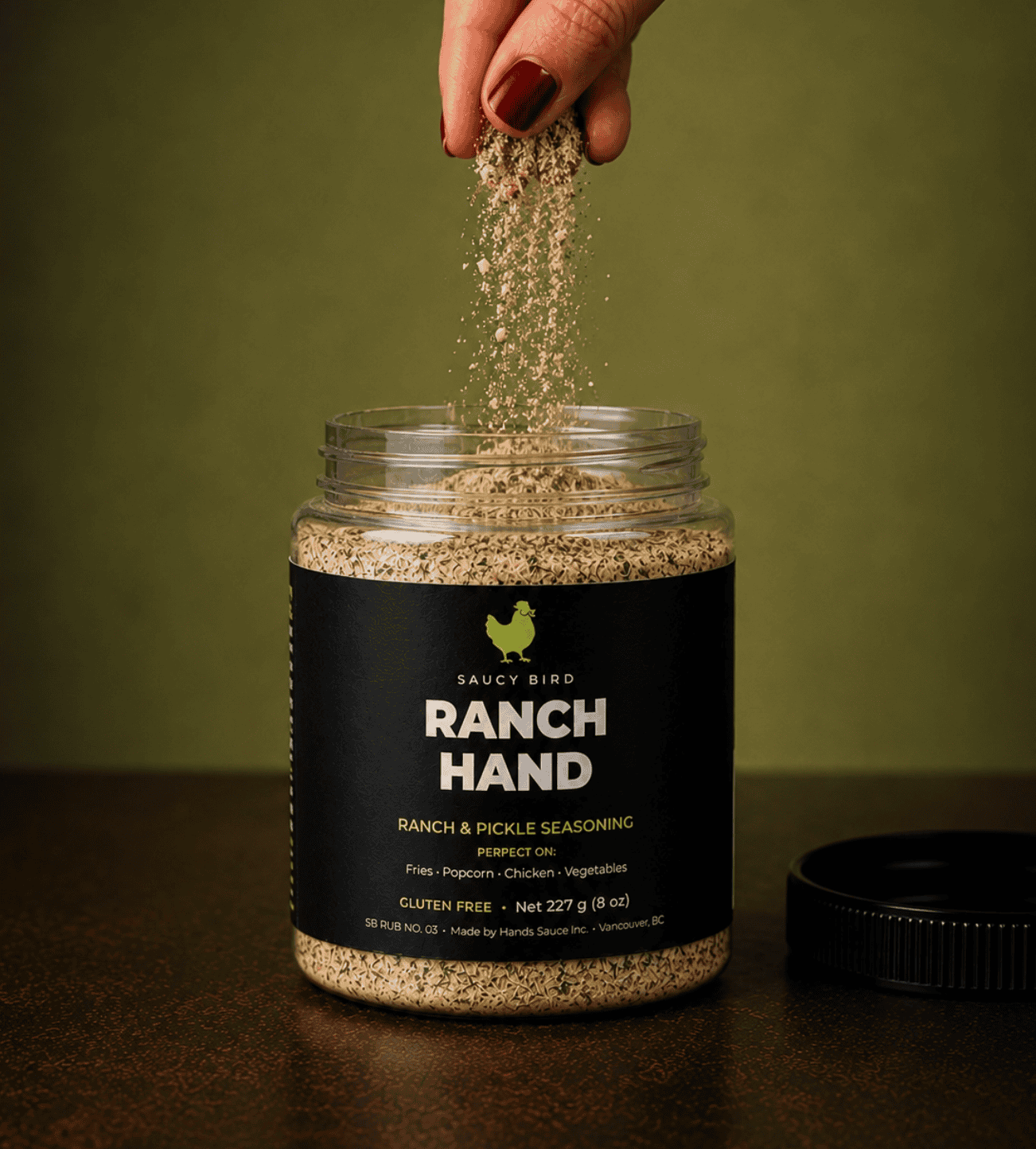 Saucy Bird Ranch Hand ranch and pickle seasoning being sprinkled from a hand into the open jar on a green background