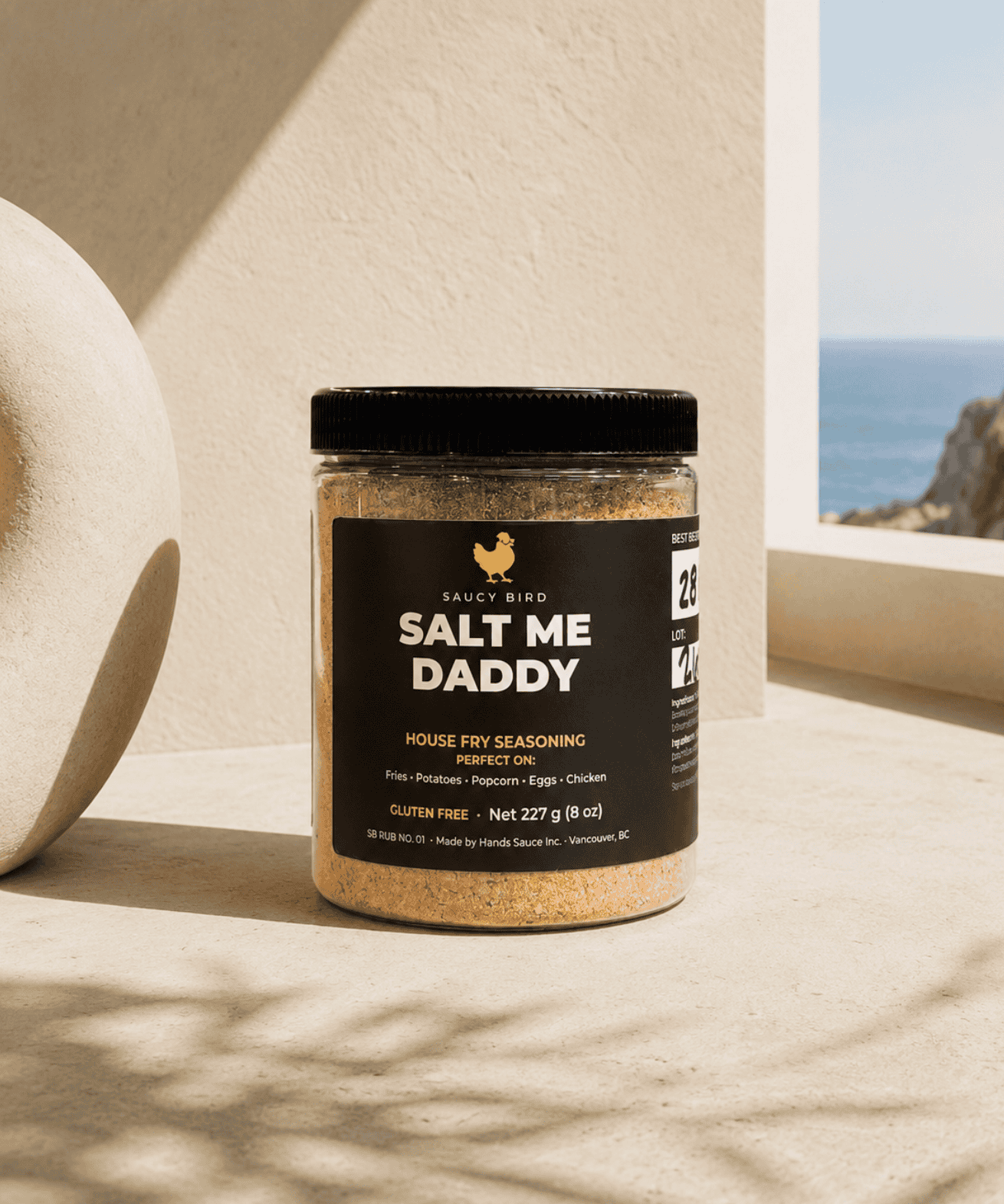 Jar of Saucy Bird Salt Me Daddy house fry seasoning on a sunlit ledge with an ocean view