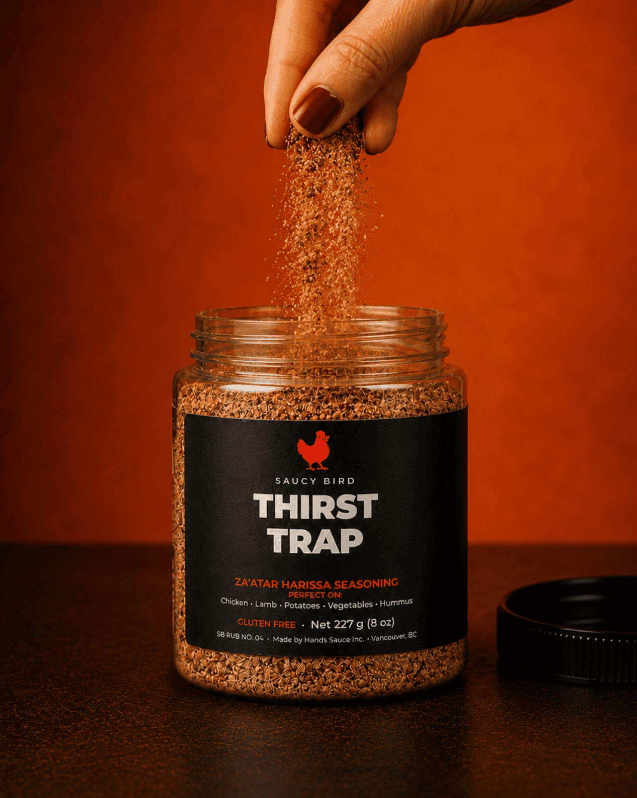 Saucy Bird Thirst Trap za'atar harissa seasoning being sprinkled from a hand into the open jar on an orange background