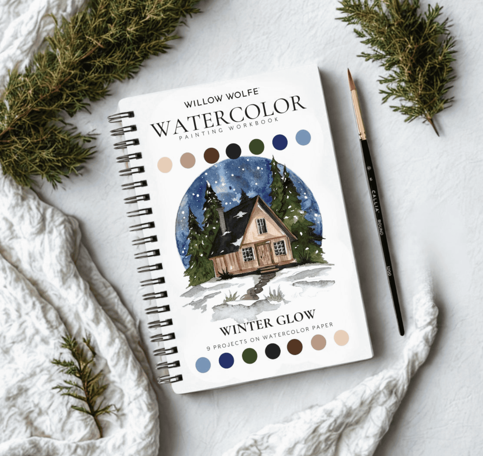 Winter Glow watercolour workbook front cover styled with evergreen sprigs and a Callia round brush